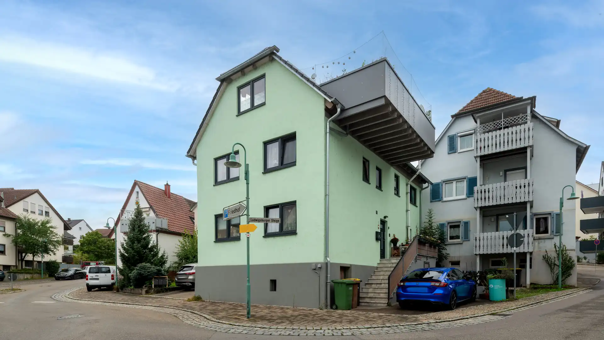 Grünes dreistöckiges Haus an der Straße gelegen mit großer grauer Dachterrasse und blauem AUto vor dem Haus